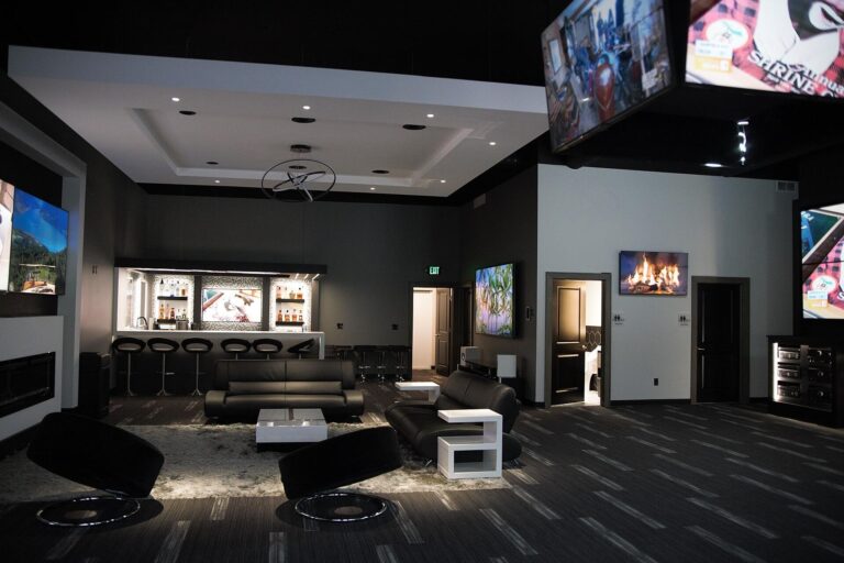 A modern lounge with a sleek black and gray interior featuring a bar area with illuminated shelves stocked with liquor bottles.   Pure AV Luxury Home Automation 768x512